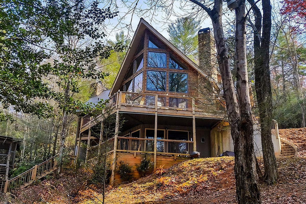 House plan exterior. Two-story A-frame style home with large windows, a wraparound deck, and a stone chimney. Built on a raised foundation.