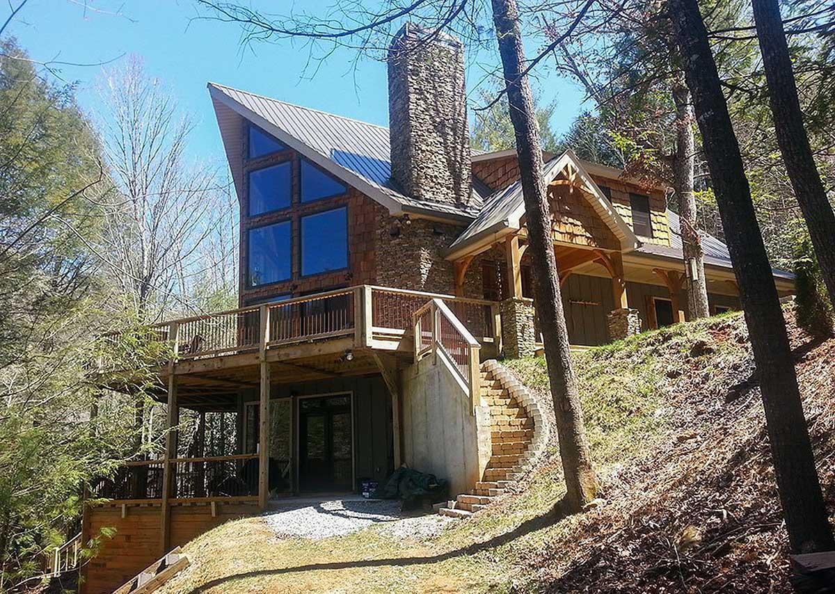 House plan exterior: Modern style with prominent stone chimney, metal roof, and large geometric window array. Features a wrap-around deck.