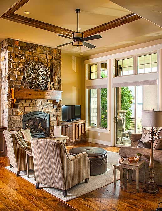 Living room with stone fireplace, wood ceiling fan, large windows, and wood floors.