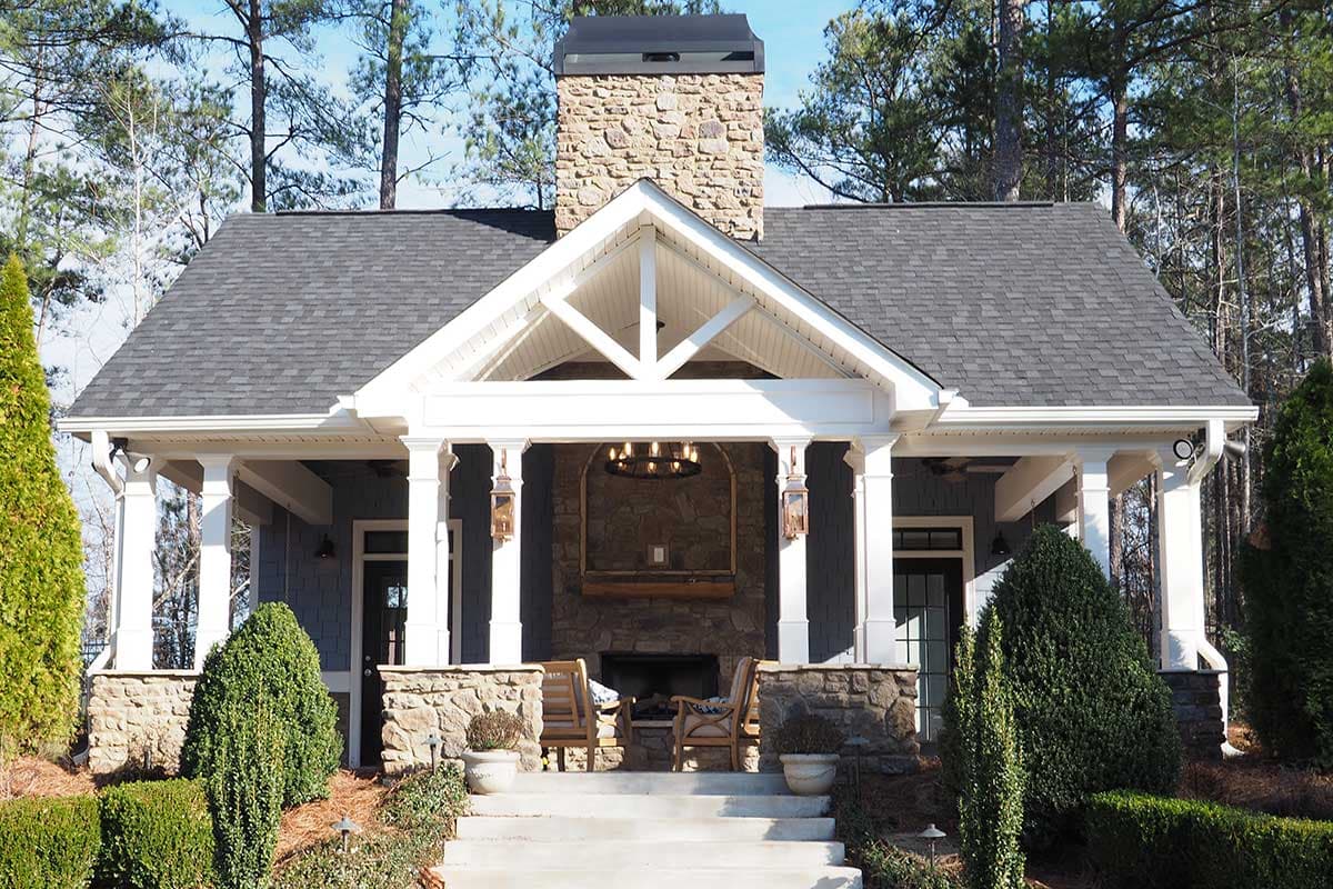 House plan exterior with covered front porch, stone chimney, exposed rafter tails, and stone foundation.