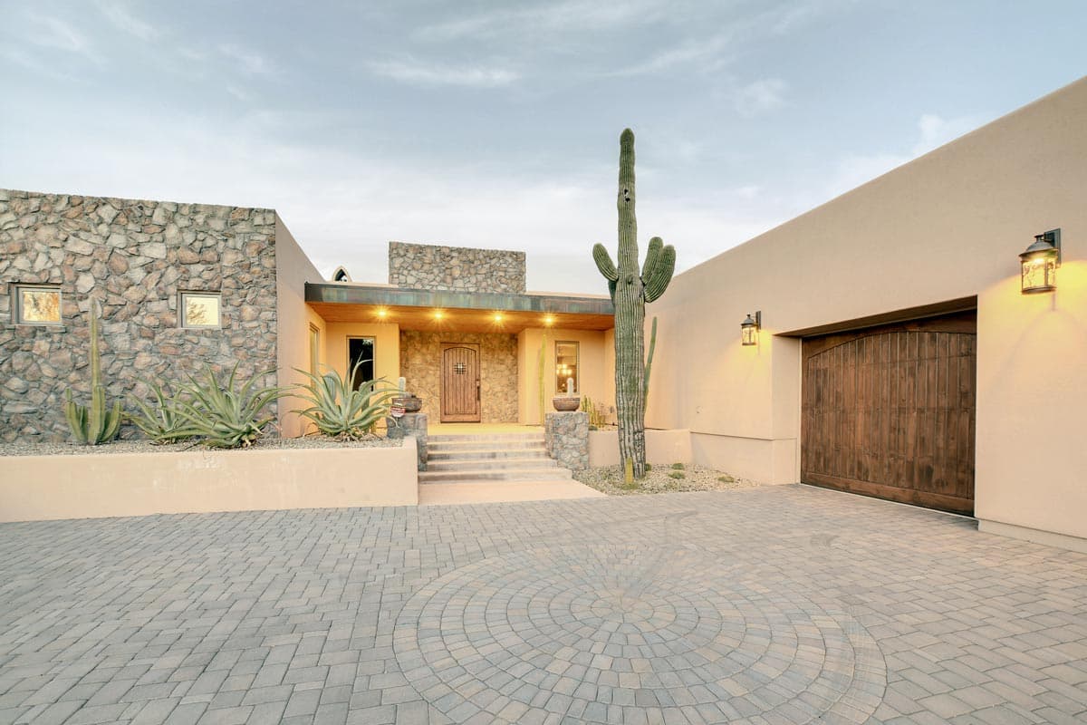 House plan exterior with stone facade, prominent saguaro cactus, wooden garage door, and paved circular driveway.