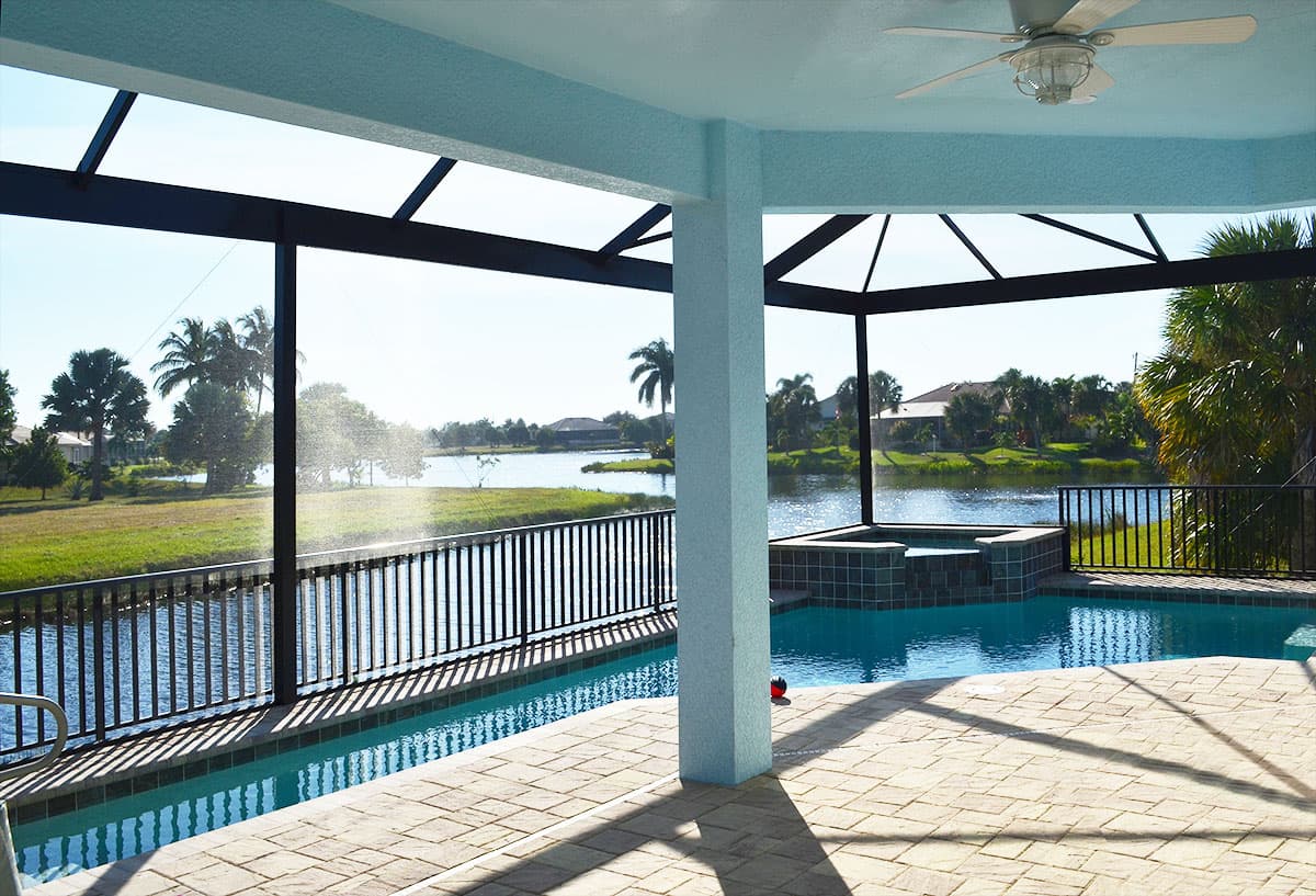 Covered patio with pool and spa overlooks a lake, featuring paver decking, black metal railing, and palm trees.