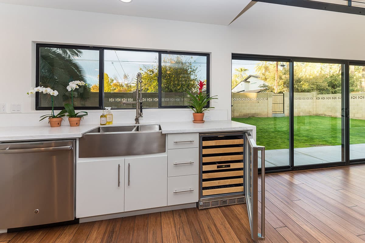 Kitchen with stainless steel farmhouse sink, wine fridge, and large windows overlooking a backyard.