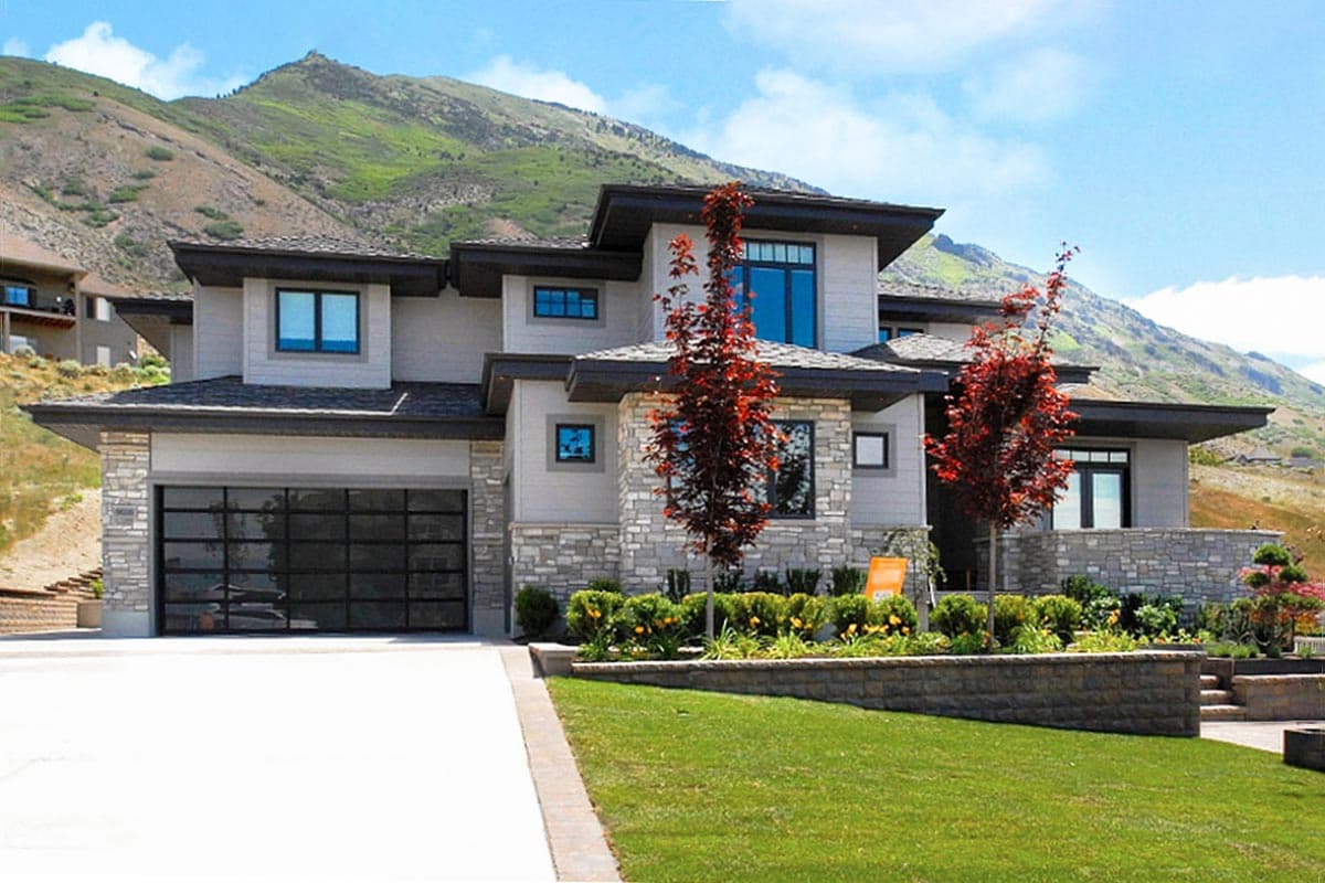 House plan exterior. Two-story, contemporary style home with flat rooflines, stone facade, and glass garage door. Landscaped front yard.