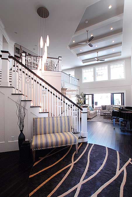 Interior view of a two-story house with a prominent staircase, coffered ceiling, and open living area with large windows.