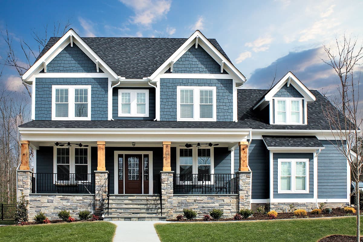 House plan exterior. Two-story Craftsman home with gabled roof, prominent front porch, dormer, and shake siding. Features stone facade and columns.