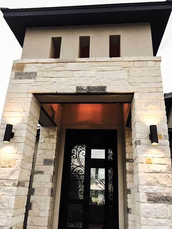 Modern house exterior with stone facade, dark metal roof, ornate double doors, and two exterior wall sconces.