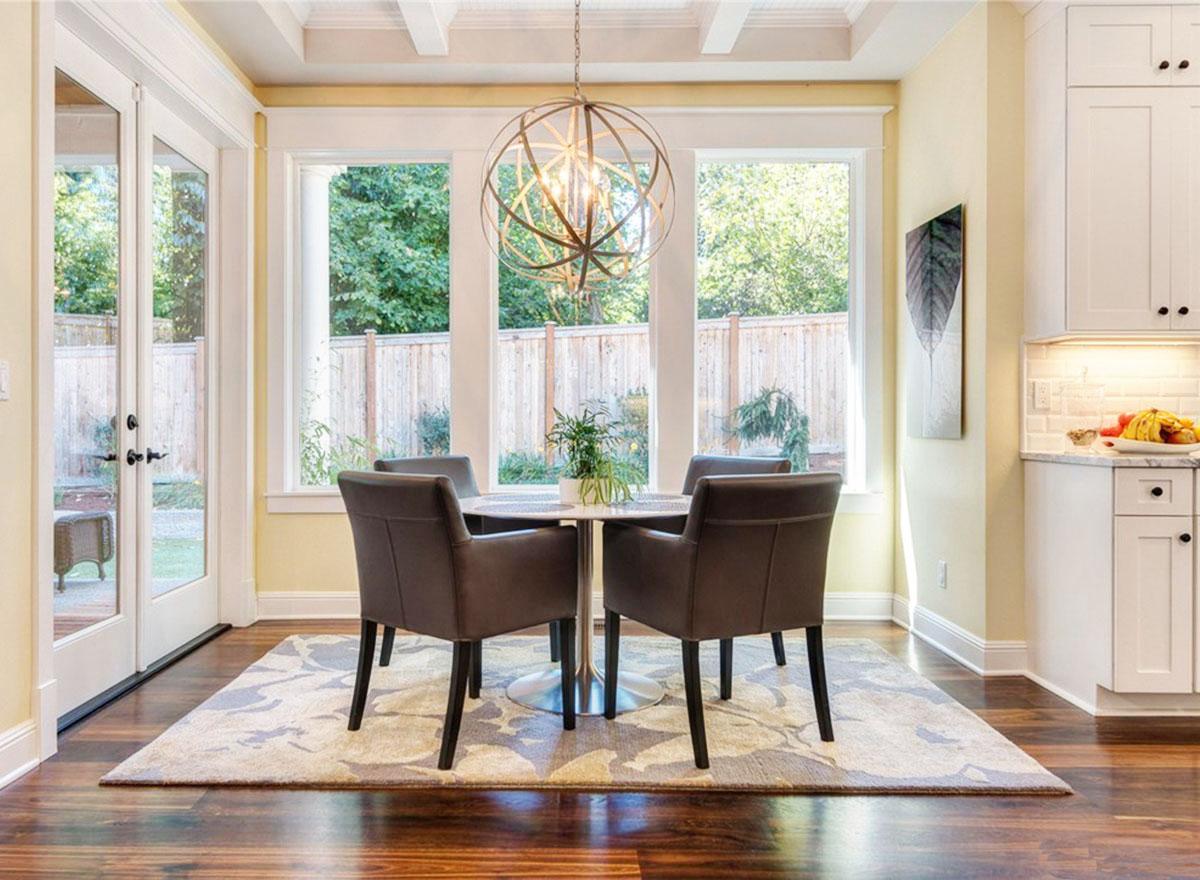 Dining area with table, four chairs, patterned rug, and large windows overlooking backyard. Adjacent kitchen features white cabinetry.