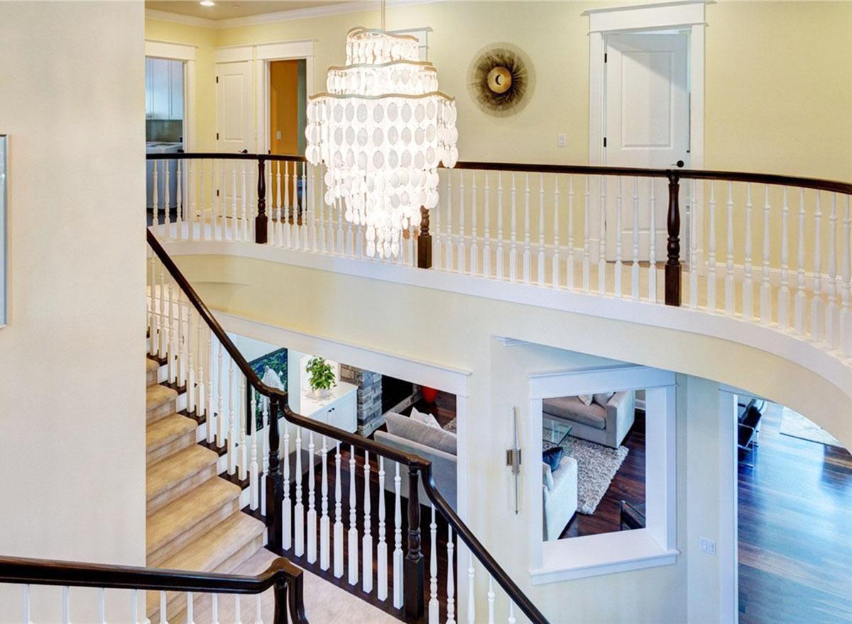Interior view of a two-story house interior with a grand staircase, white balusters, dark wood railings, and a large chandelier.