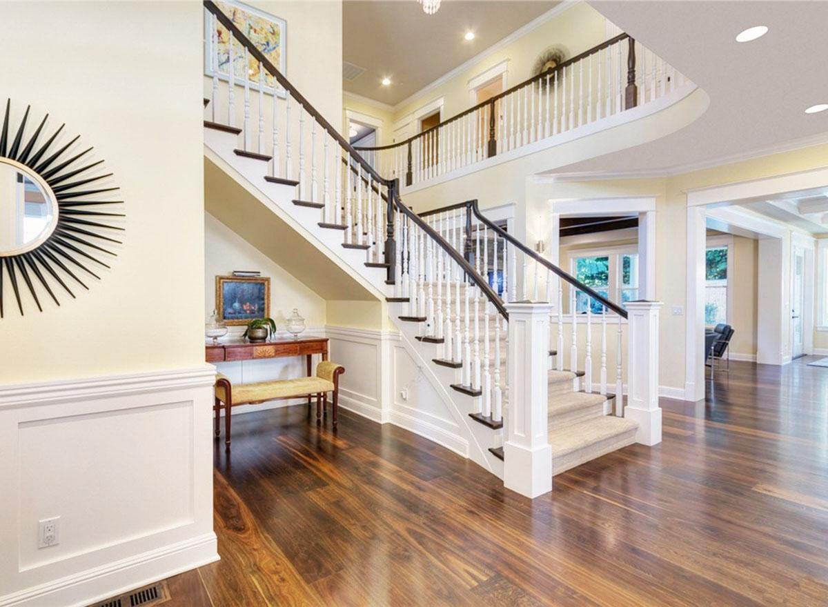 Interior view of a grand staircase with dark wood banisters, white balusters, carpeted treads, and hardwood flooring.