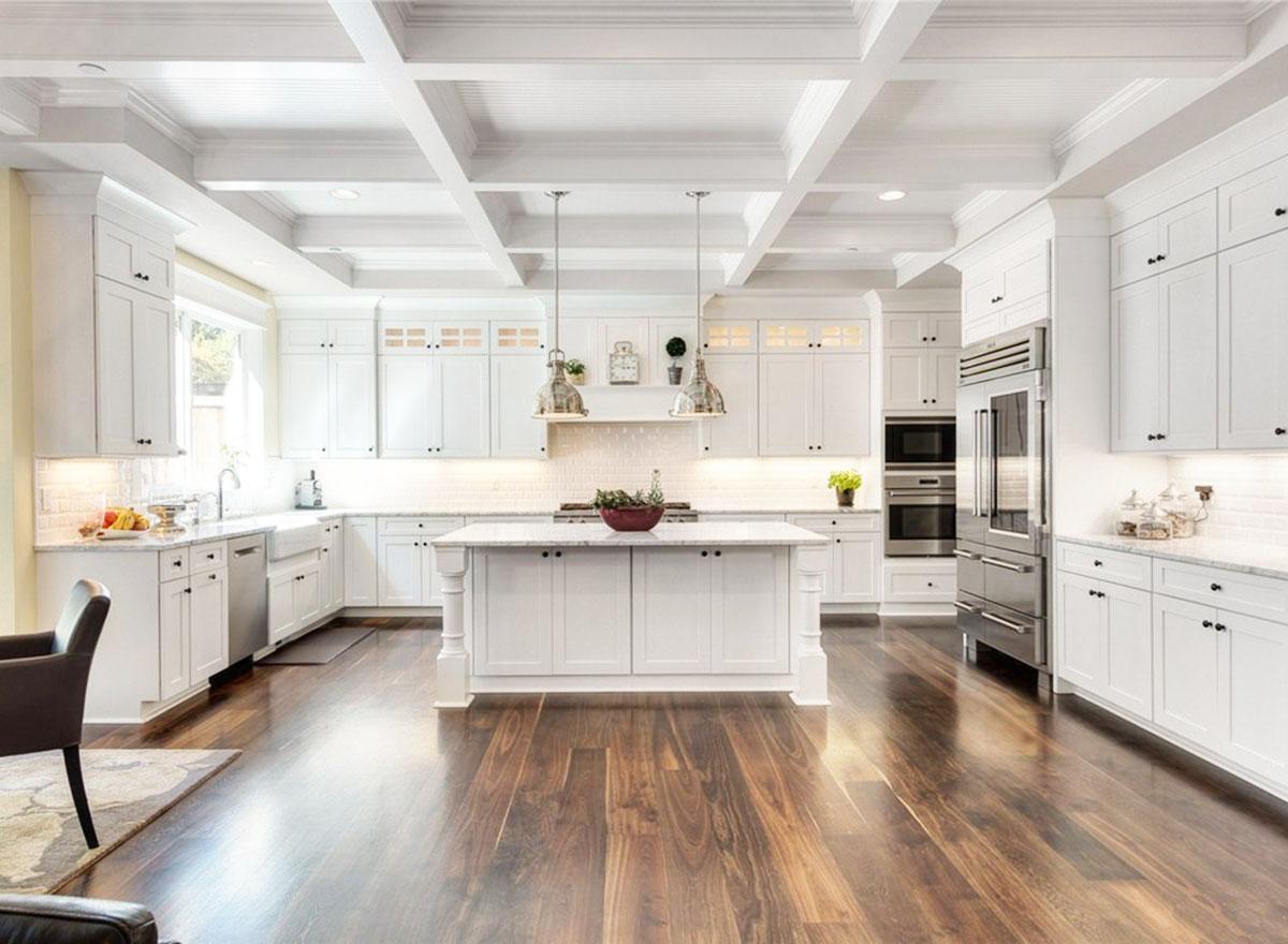 White kitchen with coffered ceiling, central island, white cabinets, marble countertops, stainless steel appliances, and hardwood floors.