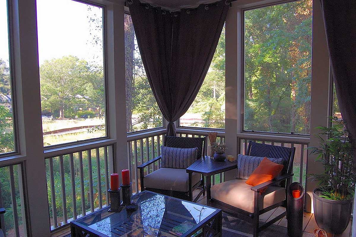 Screened porch with seating area, glass-top coffee table, and view of wooded landscape through large windows.