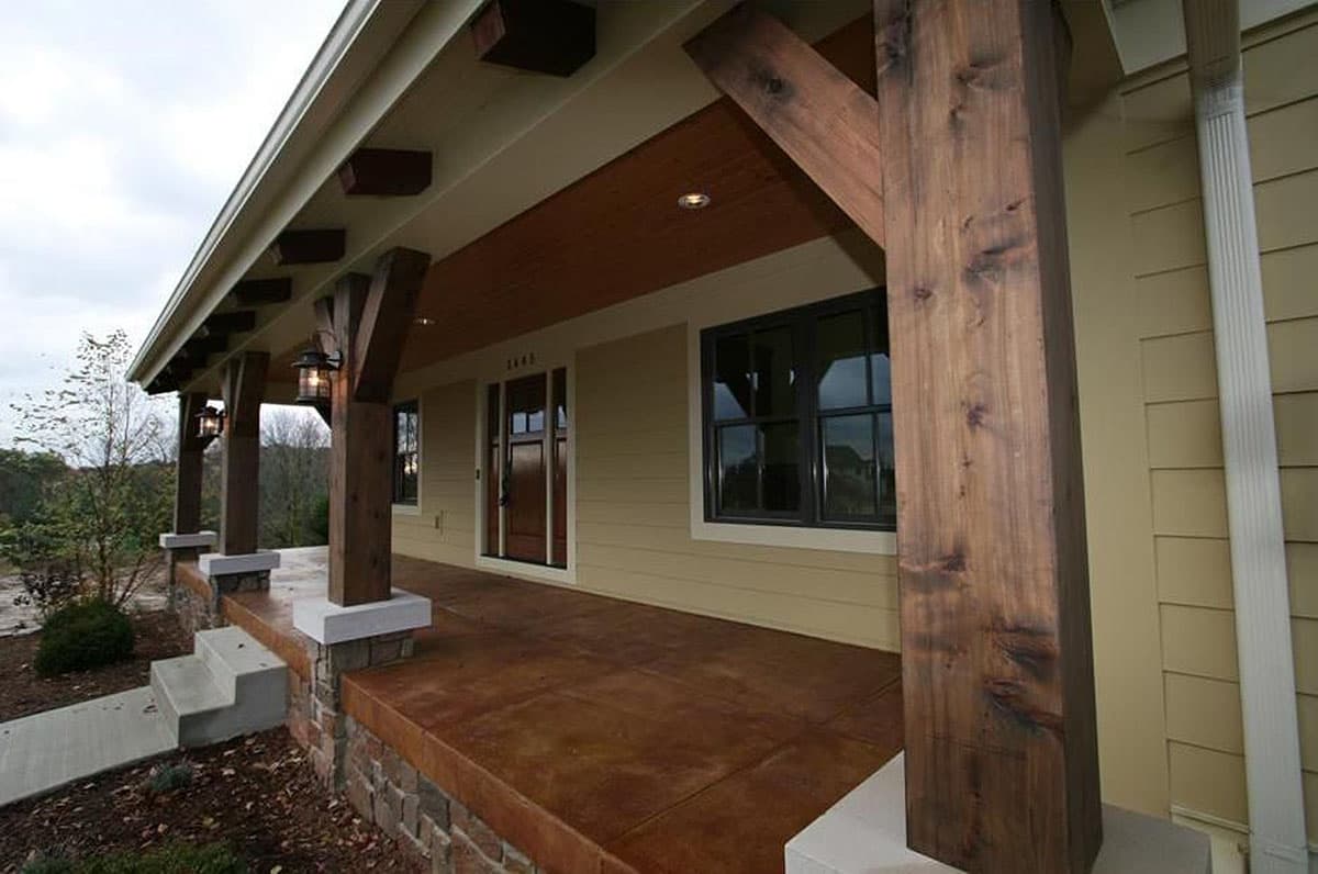 House plan exterior showing a covered porch with stained concrete, stone base, and exposed wooden beams supporting the roof.