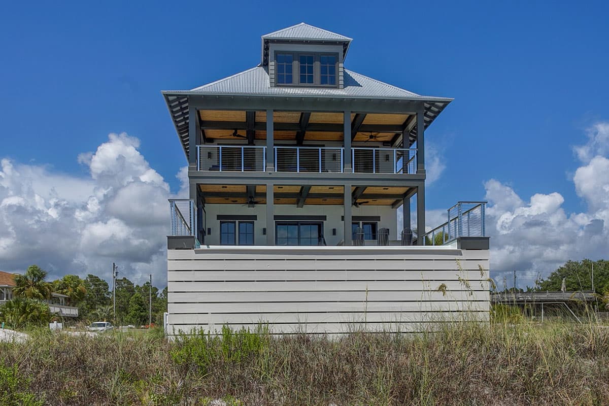 House plan exterior: Modern coastal style, three stories, metal roof, open balconies, and a raised foundation with horizontal siding.