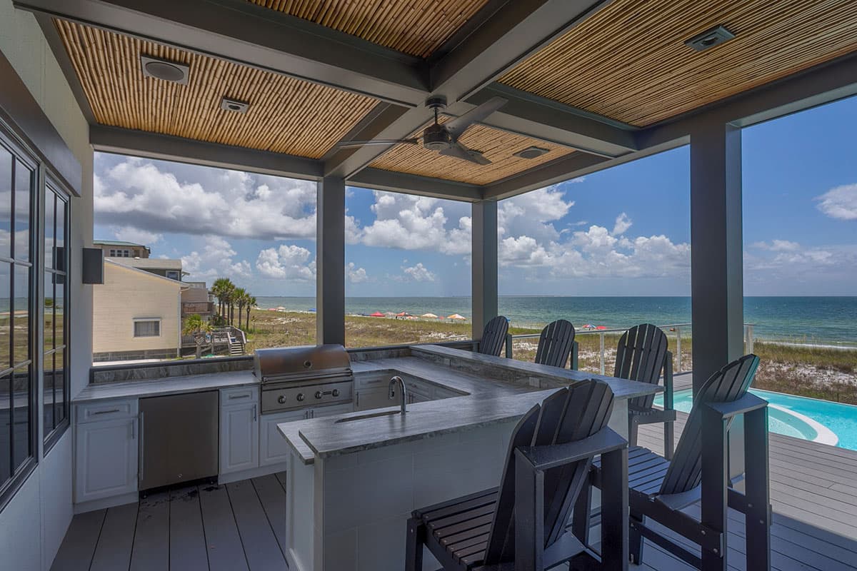 Covered outdoor kitchen with U-shaped counter and bar seating overlooks ocean. Features grill, sink, mini-fridge, fan, and bamboo ceiling.