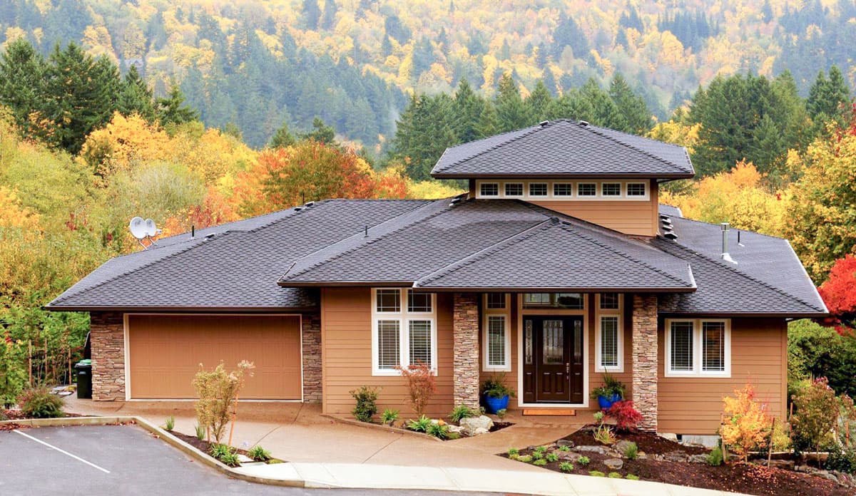 House plan exterior. One-story house with a flat roofline, garage, and gabled entry. Stone columns frame the front door. Set against autumn trees.