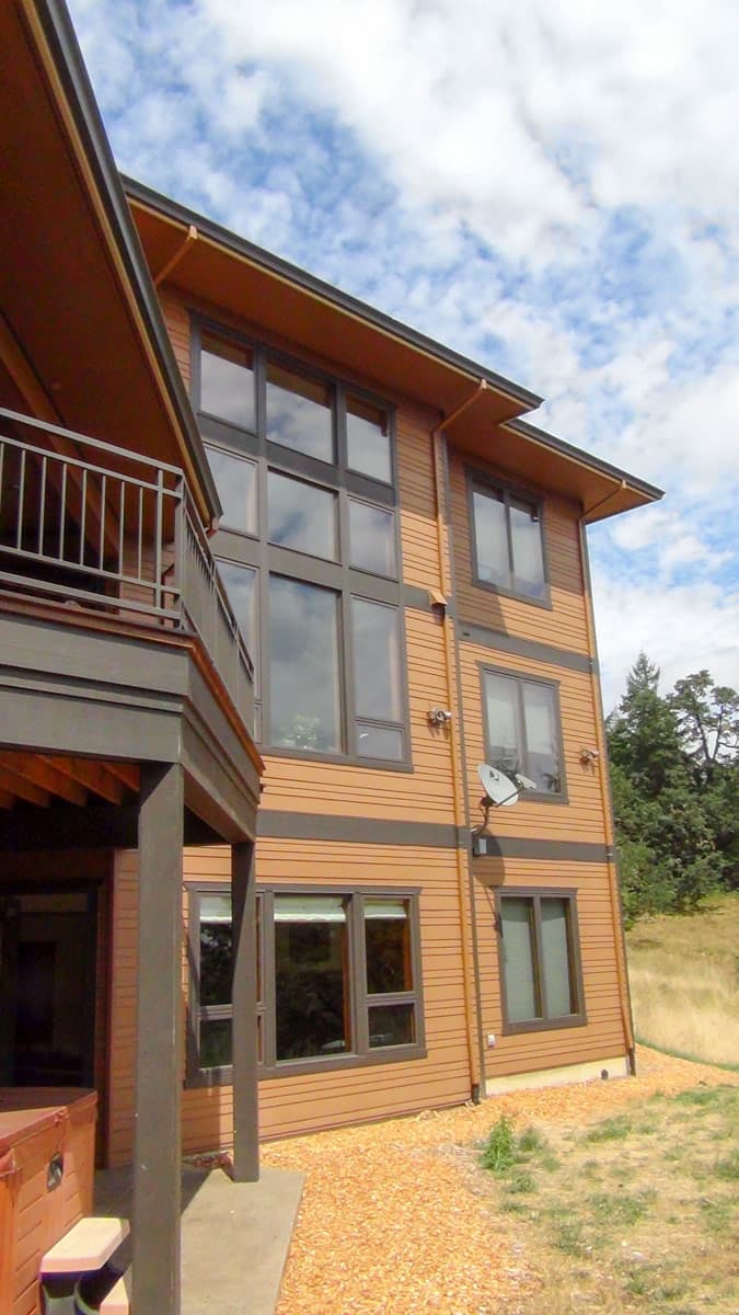 Modern house plan exterior with tan siding, dark trim, multiple stories, and large window arrangements. Features a deck with metal railing.