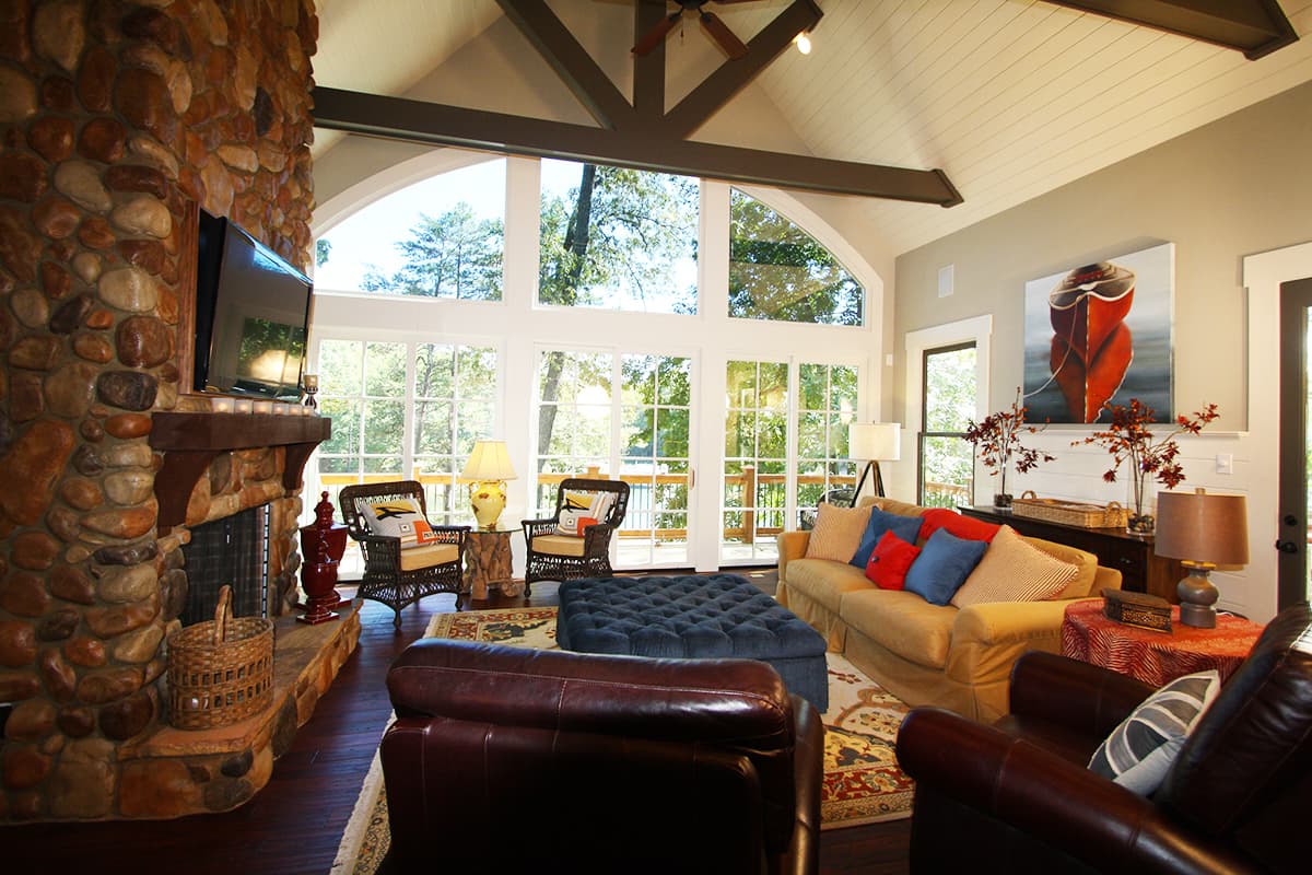 Great room interior with stone fireplace, vaulted ceiling, exposed beams, and large arched windows overlooking trees.