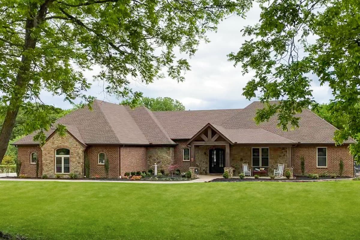 House plan exterior. One-story home with a gabled roof, stone and brick facade, and covered front porch with exposed timber beams.