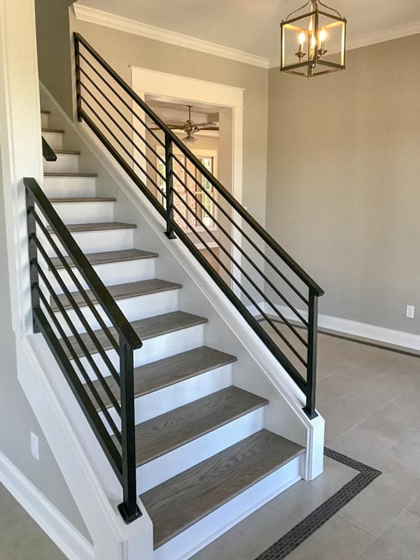 Interior staircase with white risers, gray wood treads, and black metal horizontal railing. View into a room with a ceiling fan.