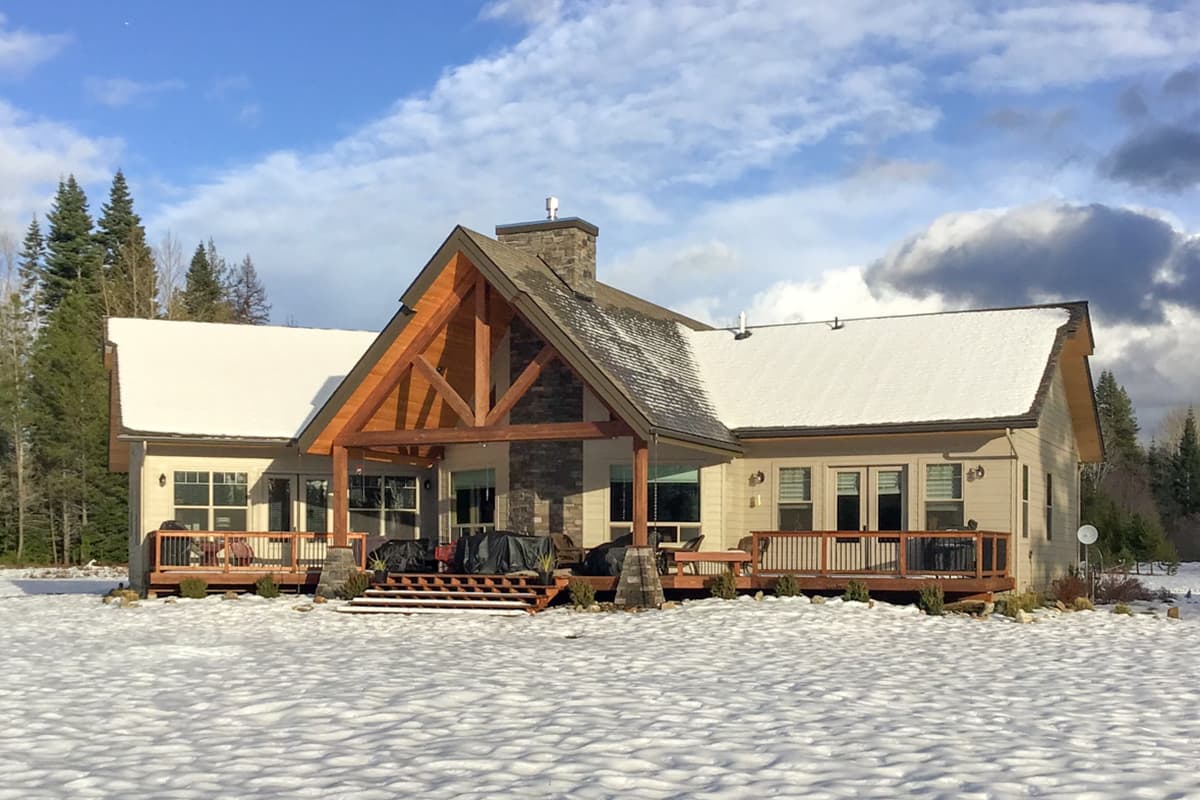 House plan exterior with exposed timber framing on a covered porch, stone chimney, and gable roof. Two-story structure with a wrap-around deck.