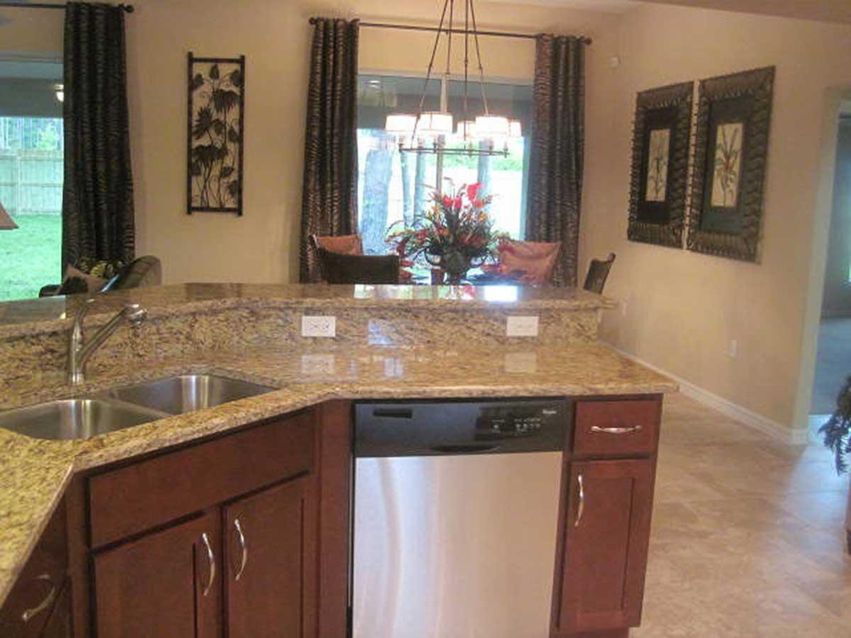 Kitchen island with granite countertop, double sink, dishwasher, and dark wood cabinets. Dining area visible beyond.