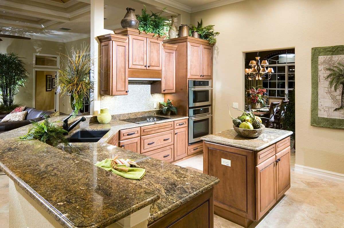 Kitchen with granite countertops, wood cabinets, double ovens, and island, opening to a dining room.