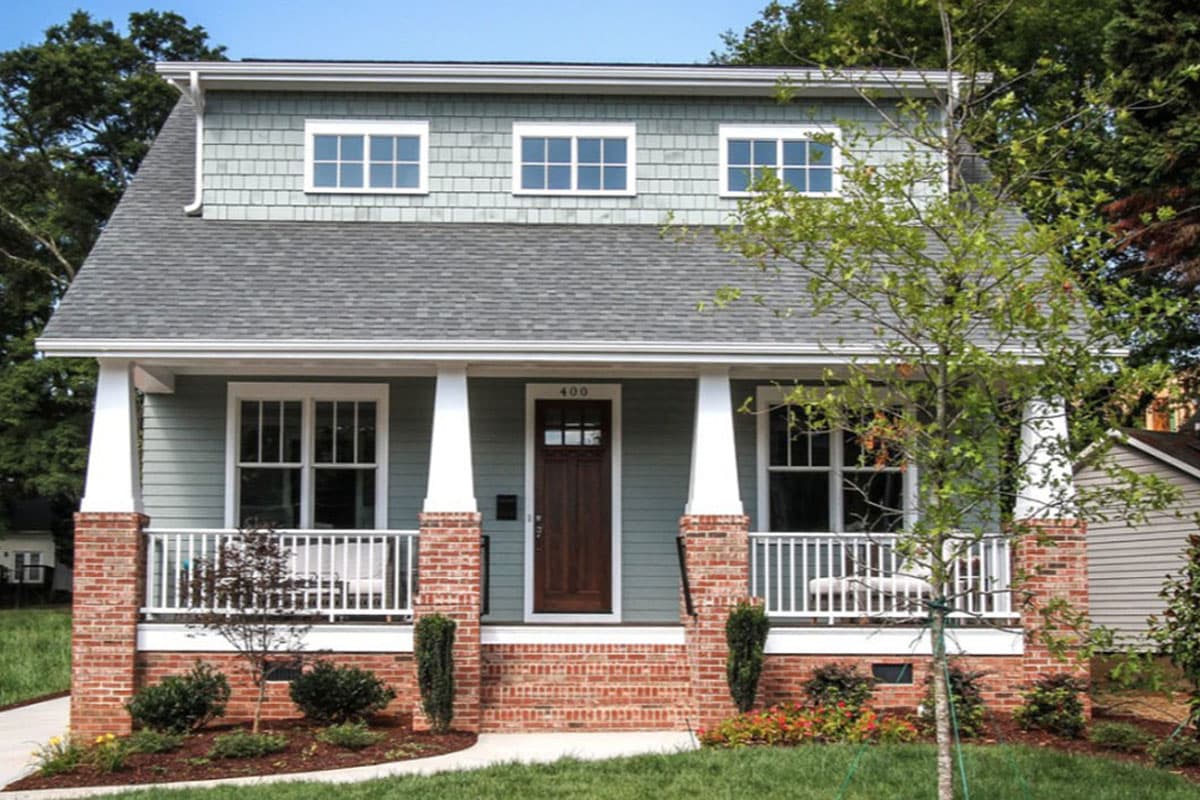 Two-story house plan exterior with covered porch, shingle siding, brick foundation, and three dormer windows.