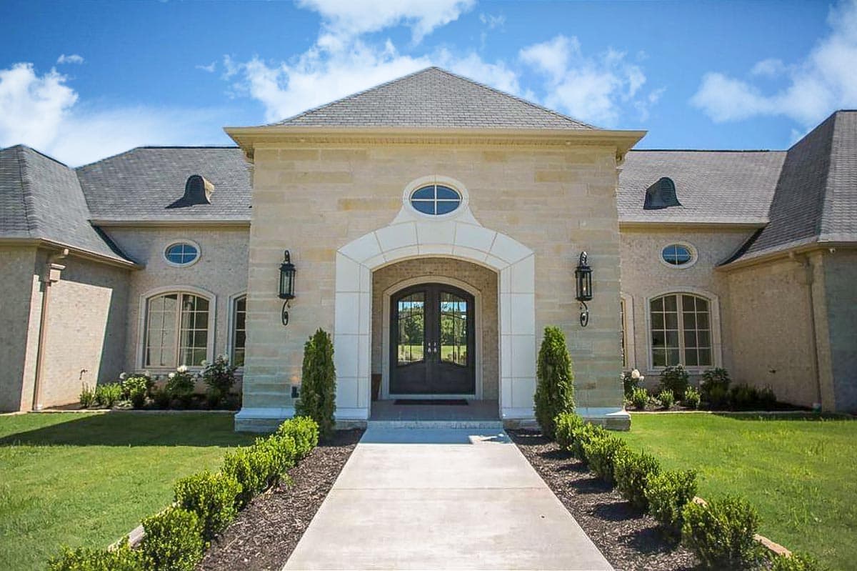European-style house plan exterior with stone facade, arched entryway, double doors, oval transom windows, and mansard roof dormers.