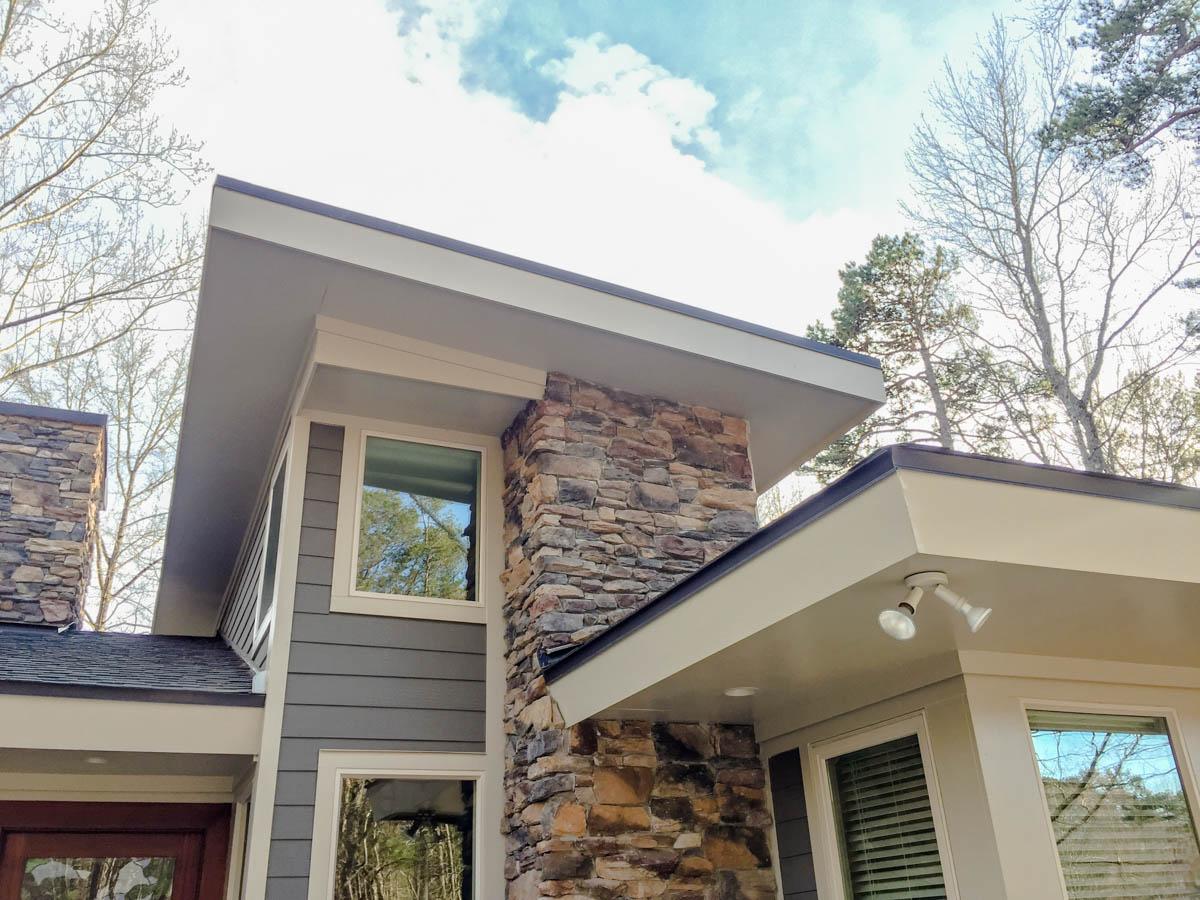 House plan exterior with stacked stone accents, horizontal siding, and intersecting shed rooflines under a cloudy sky.