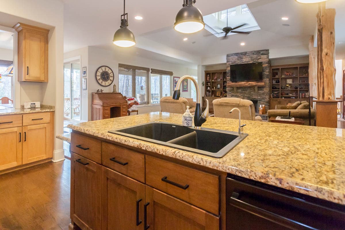 Kitchen island with double sink, granite countertop, and wooden cabinetry, opening to a living area with stone fireplace and built-in shelves.
