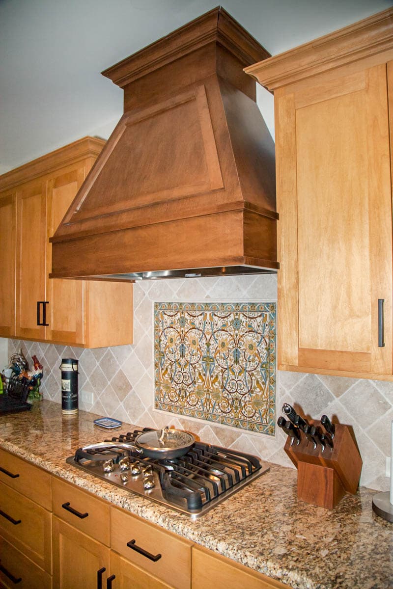 Kitchen interior showing a gas range, granite countertop, wooden cabinetry, decorative tile backsplash, and custom wood range hood.