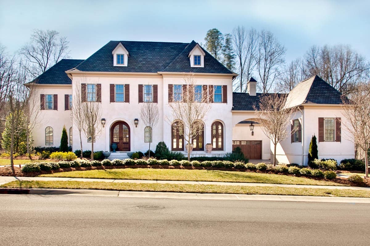 House plan exterior. Two-story home with arched entryway, dormers, and attached garage. Features dark roof and shutters.