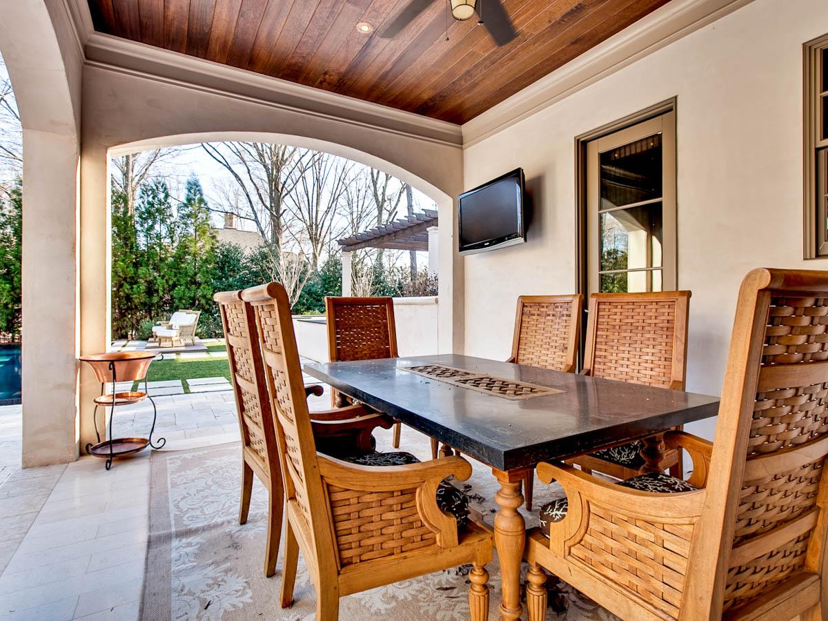 Covered outdoor dining area with stone table, wood chairs, ceiling fan, and mounted television.