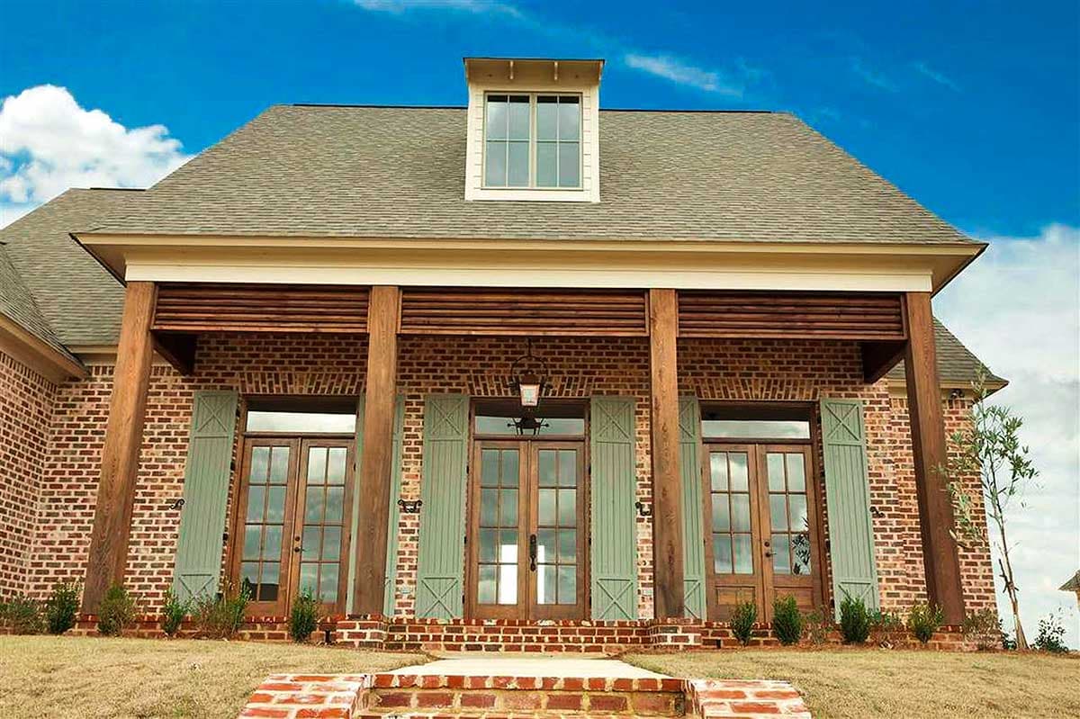 House plan exterior: Brick facade with a covered porch, exposed wooden posts, French doors, and decorative shutters. Dormer window on roof.