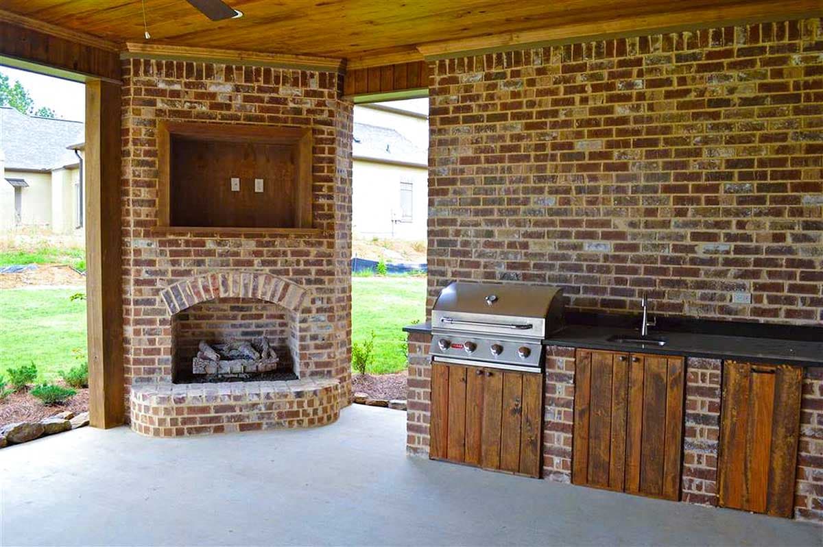 Covered outdoor living area with brick fireplace, built-in grill, sink, and wood-paneled cabinetry under a wood ceiling.