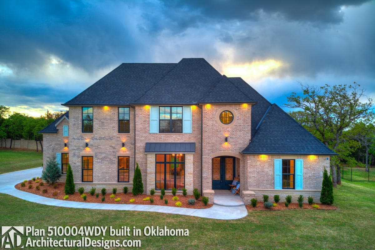 House plan exterior: Two-story brick home with a prominent gable roof, circular window, covered entry, and light blue shutters.