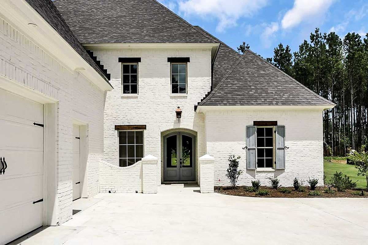 House plan exterior: White brick two-story home with arched double doors, varied window styles, and a side-entry garage.