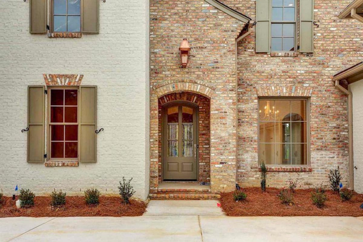 House plan exterior showing brick and white painted walls, arched entry with glass-paned door, and multi-paned windows with shutters.