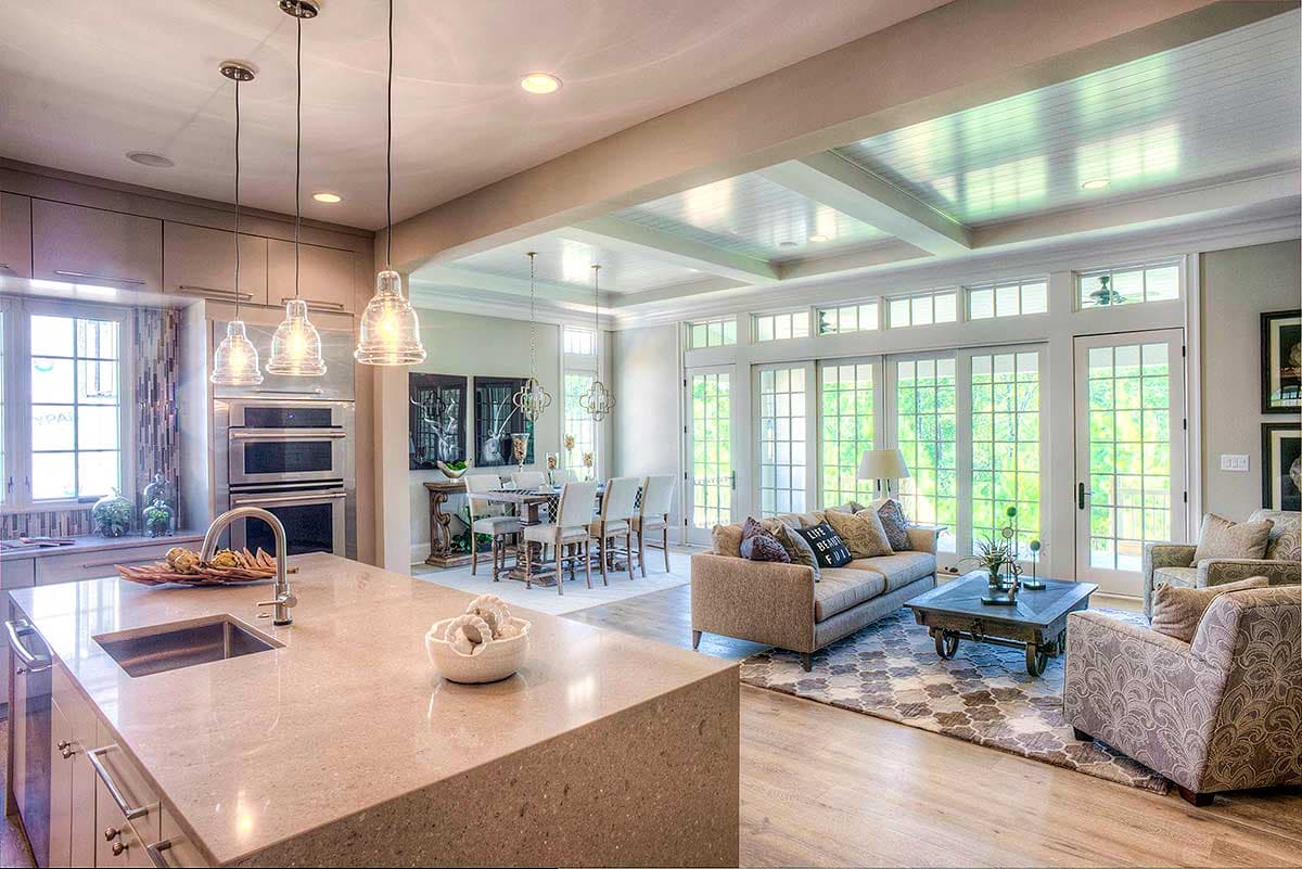 Interior view showing a kitchen island, dining area with table and chairs, and a living room with sofas, large windows, and a coffered ceiling.