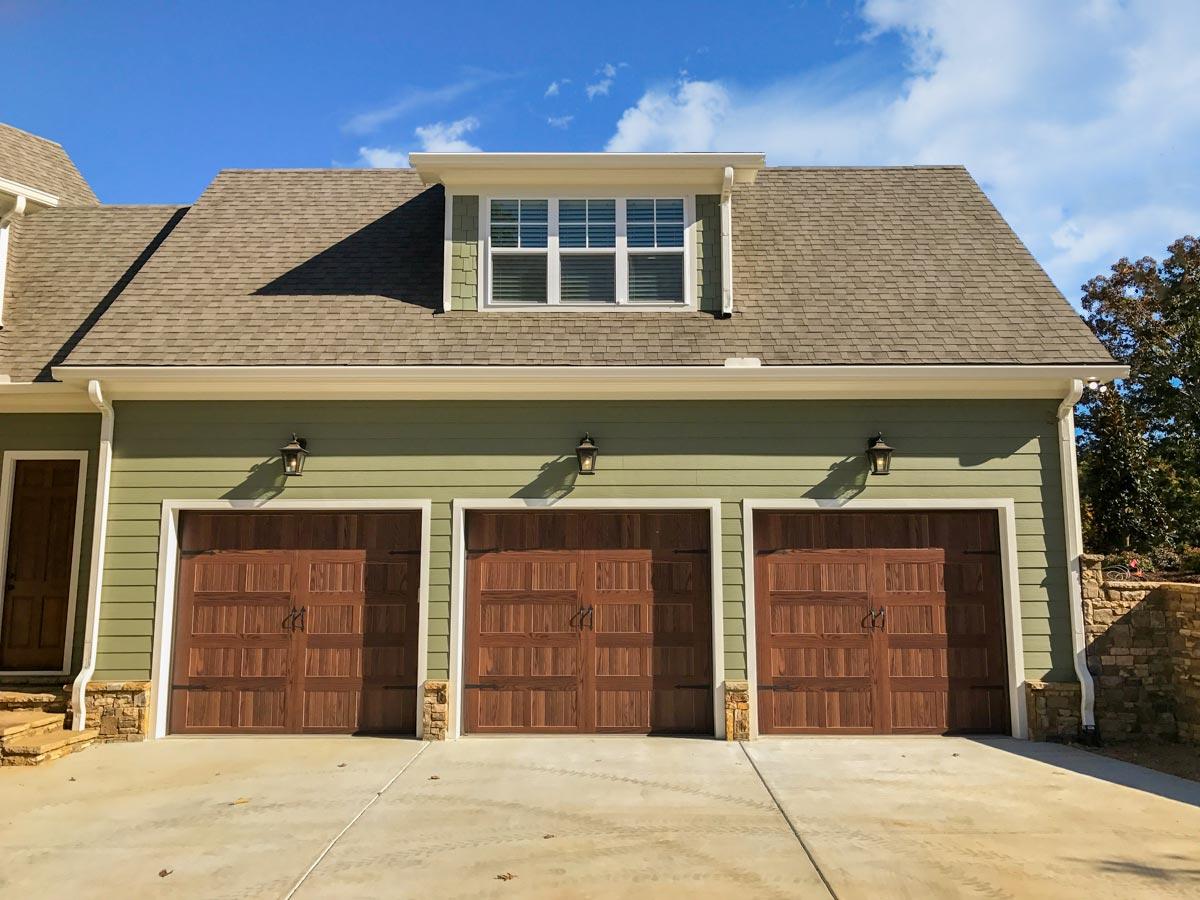 House plan exterior: Three-car garage with wood doors, green siding, dormer window, and shingle roof.