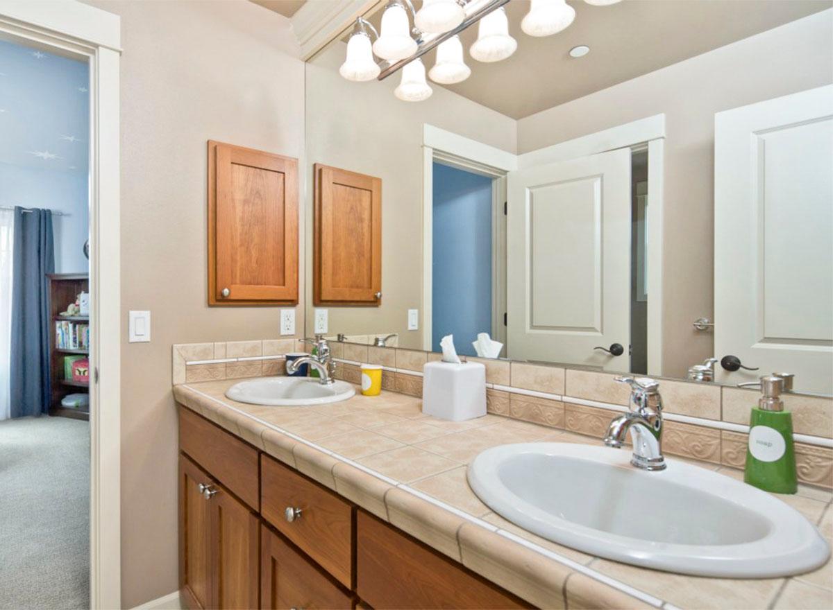 Double vanity bathroom with two sinks, wood cabinetry, tiled backsplash, and dual overhead cabinets. View of adjacent room with bookshelf.