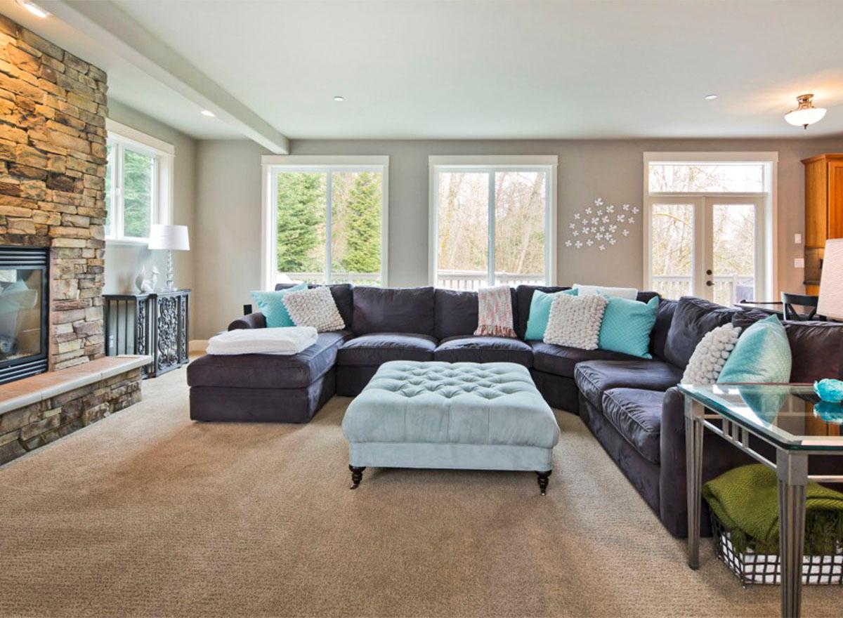 Living room with stone fireplace, large sectional sofa, ottoman, and multiple windows with wooded view.