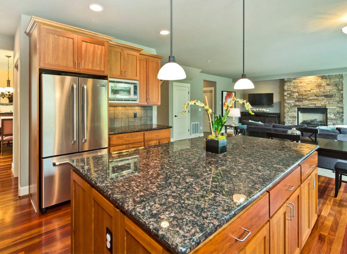 Kitchen with island, stainless steel refrigerator, wood cabinetry, and view into living room with stone fireplace.