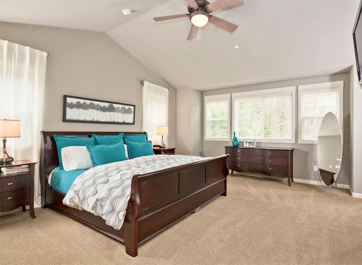 Bedroom with vaulted ceiling, wood bed frame, dresser, and three large windows.