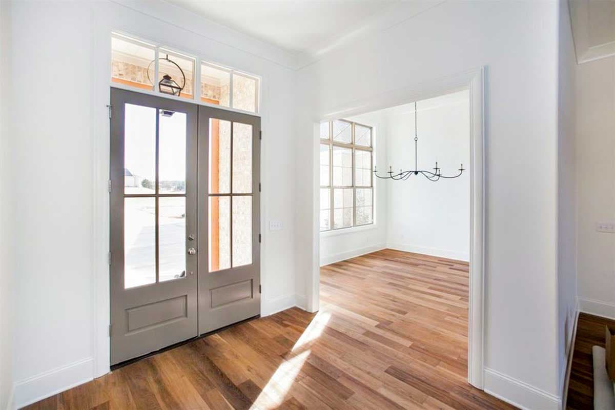 Interior view of double entry doors with glass panes and transom windows, opening to a room with hardwood floors and a chandelier.