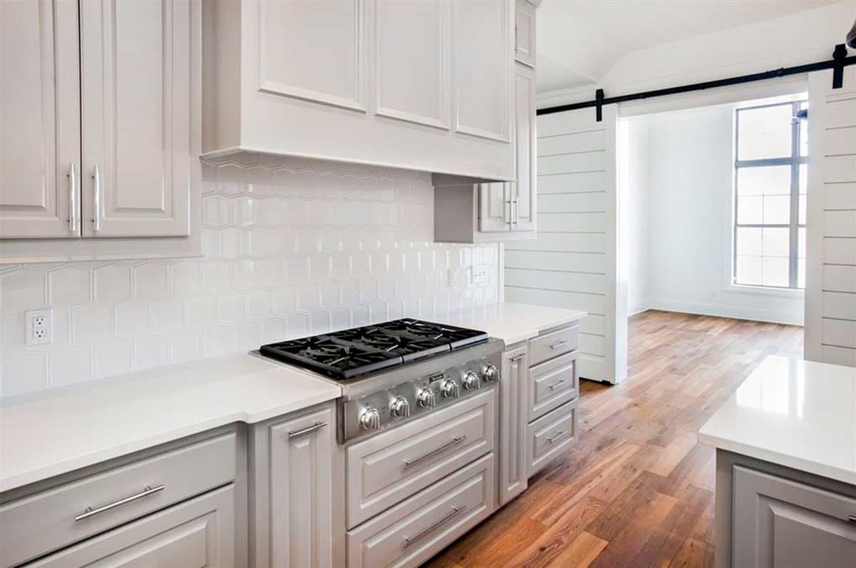 Kitchen with stainless steel gas range, white hexagonal tile backsplash, gray shaker cabinets, and sliding barn door leading to room with wood floors.