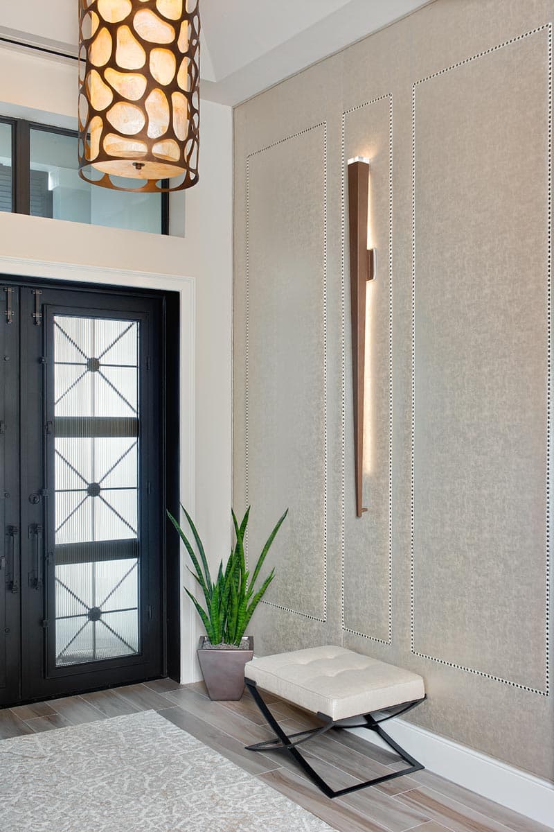 Entryway interior with double black doors, patterned glass, textured wall panels, modern lighting, and a bench.