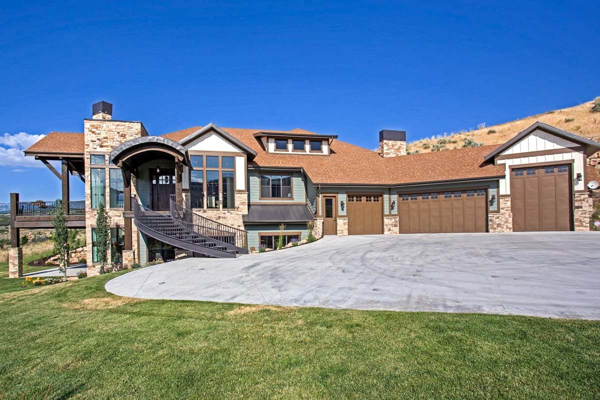 House plan exterior with a multi-level design, prominent stone chimney, curved entrance canopy, spiral staircase, and a five-car garage.