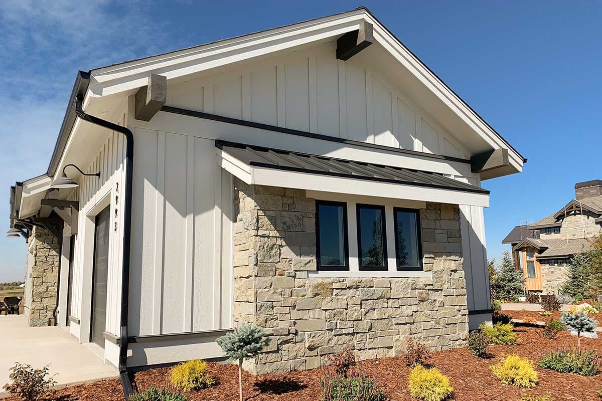 House plan exterior with white board and batten siding, stone wainscoting, metal roof awning, and exposed rafter tails.